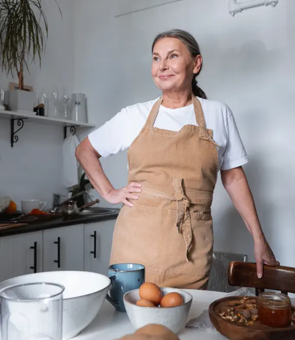 Bienfaits de la cuisine chez les personnes âgées Une femme d'une soixantaine d'année debout dans sa cuisine prête à réaliser une recette. Sur une table devant elle, des ingrédients et accessoires de cuisine.