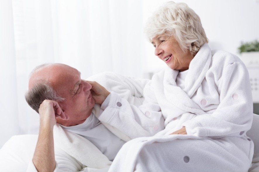 Le sexe durant la vieillesse Un couple de séniors en robes de chambre blanches qui se regardent dans les yeux en souriant. L'homme est allongé sur le coté et la femme assise contre lui, lui caresse le visage.